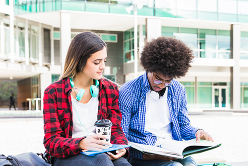 A female student in a red plaid flannel shirt holds a paper coffee cup. She sits outside next to a young man in a checkered blue button down shirt who is looking down at a textbook he is holding.
